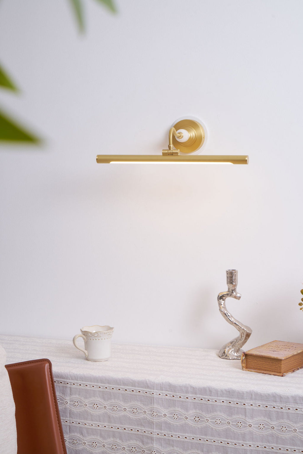 This Clérachic wall sconce in gold glows above a table set with a jug, candlestick and book, beside a leather chair.