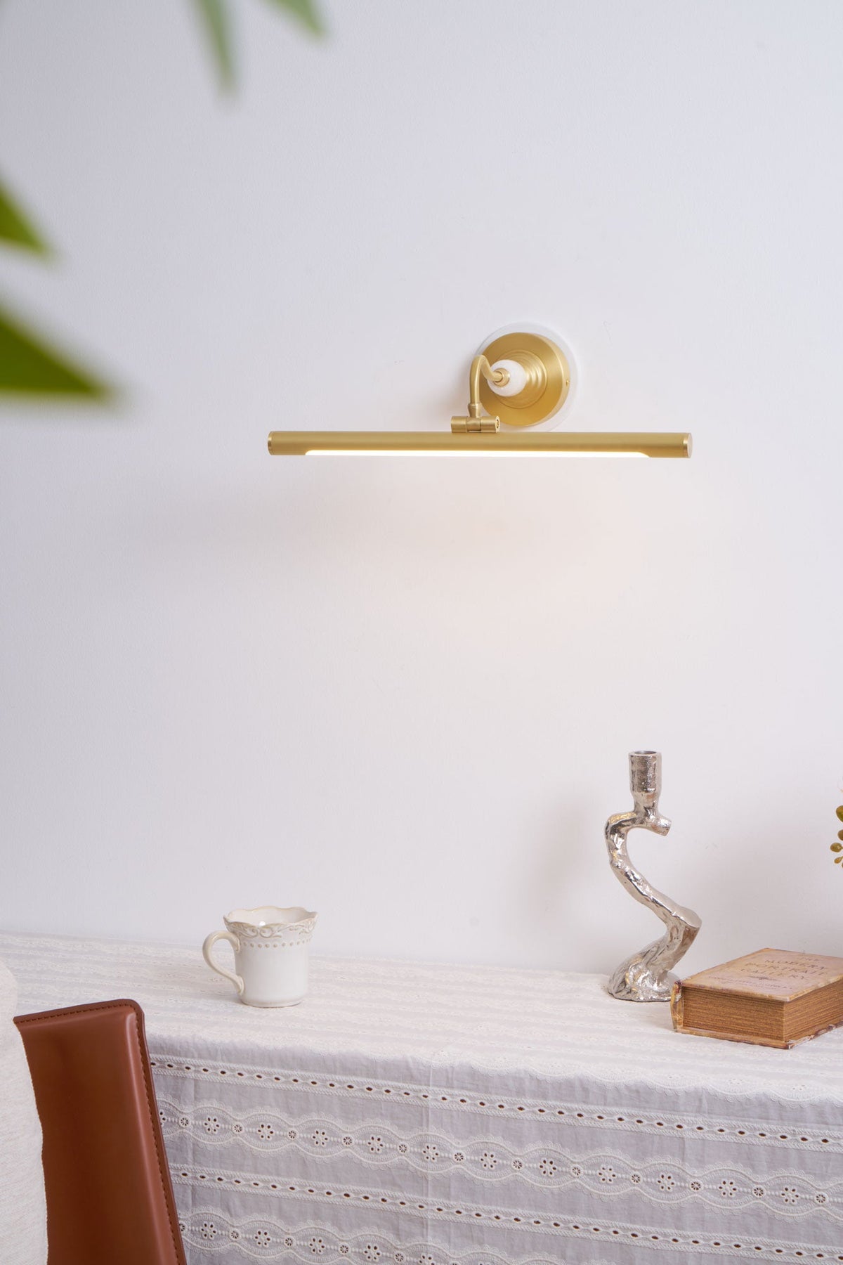 This Clérachic wall sconce in gold glows above a table set with a jug, candlestick and book, beside a leather chair.