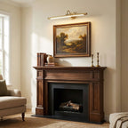 A landscape painting above a wooden fireplace mantel sits in the warm wash of this gold Clérachic wall sconce.