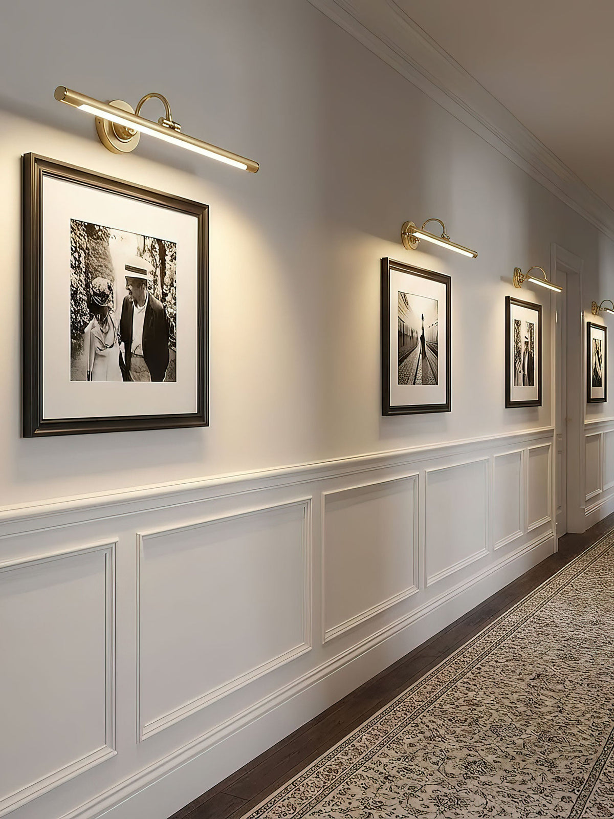 Warm light from this gold Clérachic wall sconce washes over a row of framed photographs along a wainscoted hallway.