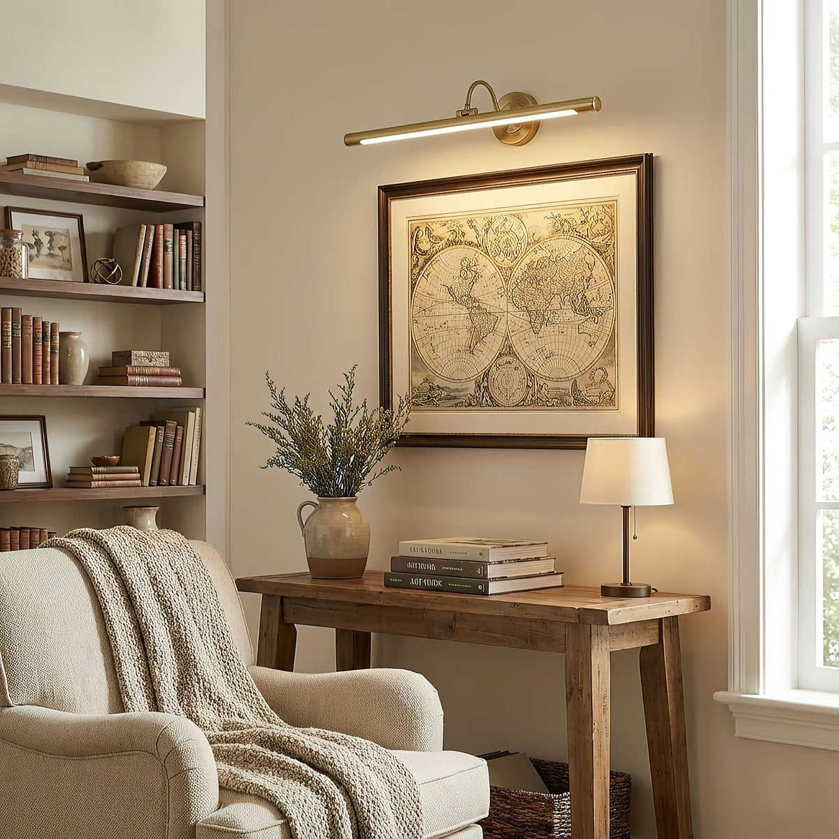 A framed map above a wooden console table is lit by this Clérachic wall sconce in gold, beside a bookshelf and armchair.