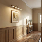 In a hallway with wood wainscoting and a patterned runner, this gold Clérachic wall sconce lights a framed artwork.