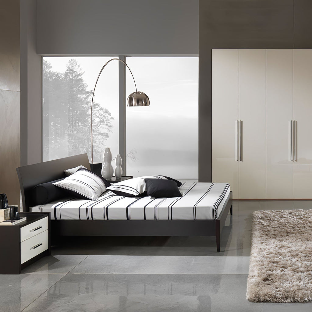 The Clérachic arc lamp curves beside a dark platform bed dressed in striped bedding, facing a window with bare winter trees.