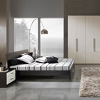 The Clérachic arc lamp curves beside a dark platform bed dressed in striped bedding, facing a window with bare winter trees.