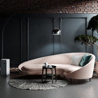 Against a dark panelled accent wall, the Clérachic arc lamp curves above a blush curved sofa and a potted olive tree.