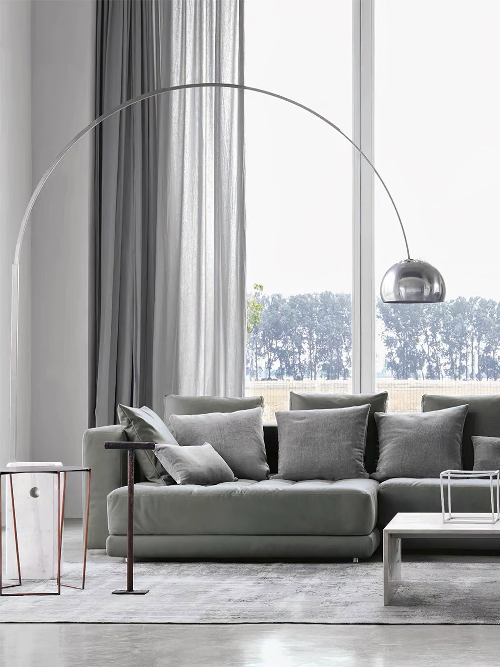 The Clérachic arc lamp's marble base sits beside a grey sectional sofa, its chrome dome framed by a tall garden-view window.