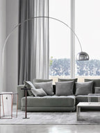 The Clérachic arc lamp's marble base sits beside a grey sectional sofa, its chrome dome framed by a tall garden-view window.