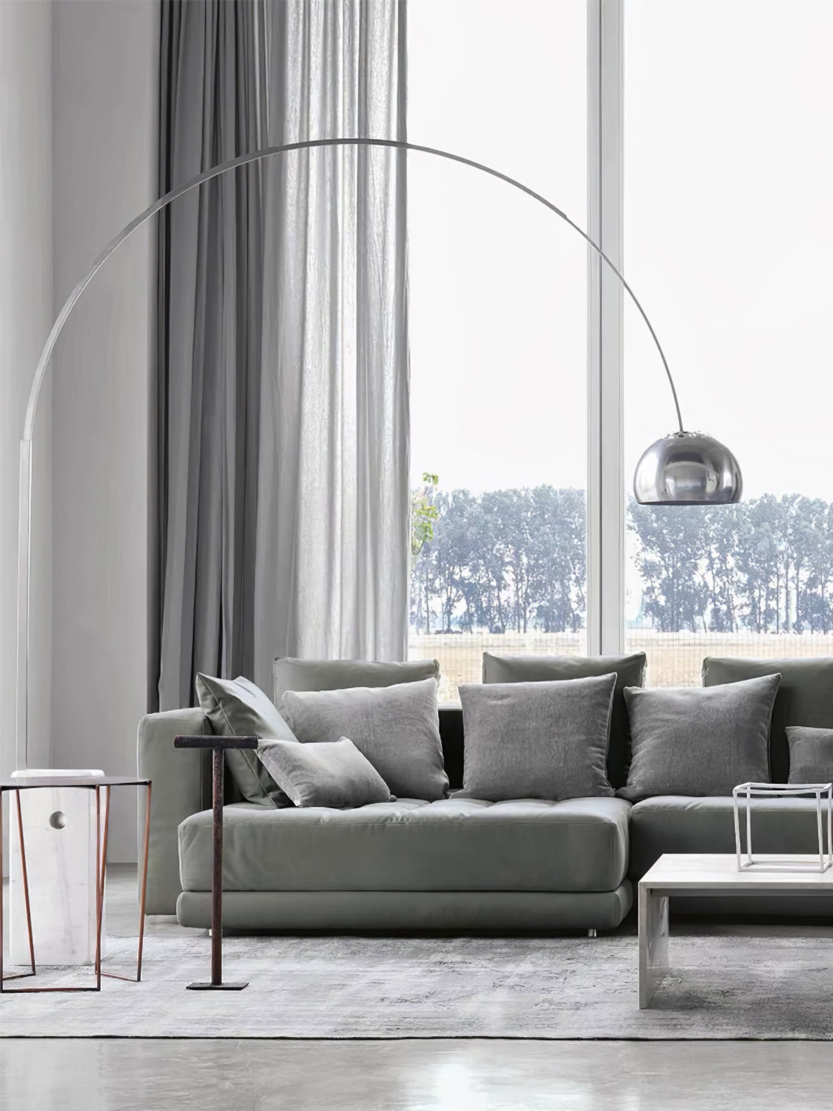 The Clérachic arc lamp's marble base sits beside a grey sectional sofa, its chrome dome framed by a tall garden-view window.