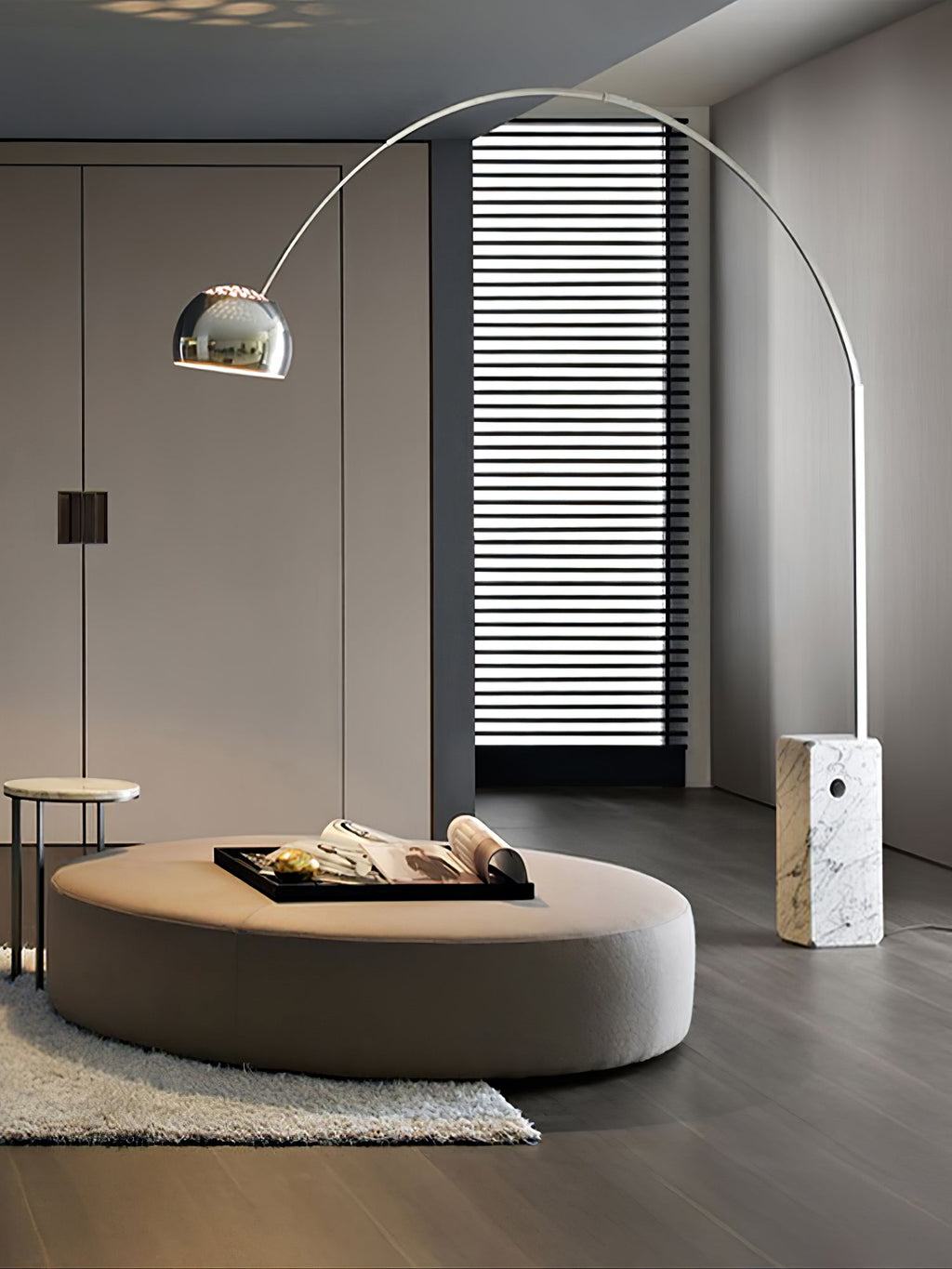 The Clérachic arc lamp's chrome dome hovers over a round taupe ottoman with a book-laden tray, light striping the wall behind.