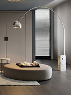 The Clérachic arc lamp's chrome dome hovers over a round taupe ottoman with a book-laden tray, light striping the wall behind.