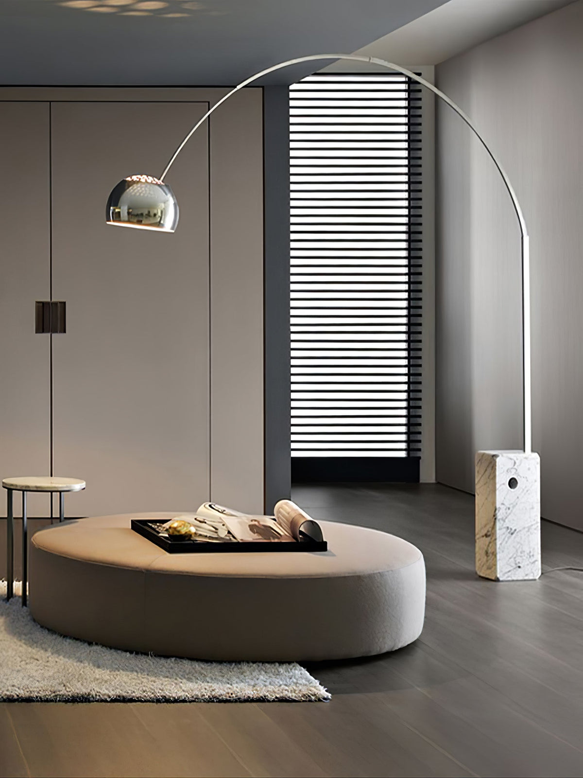 The Clérachic arc lamp's chrome dome hovers over a round taupe ottoman with a book-laden tray, light striping the wall behind.