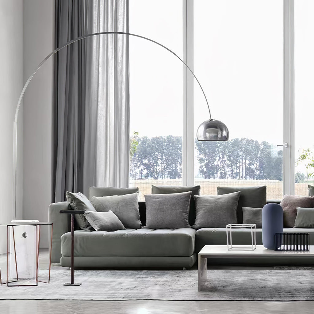 The Clérachic arc lamp's chrome dome curves above a grey corner sofa, its marble base beside a slim wire-frame side table.