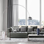 The Clérachic arc lamp's chrome dome curves above a grey corner sofa, its marble base beside a slim wire-frame side table.