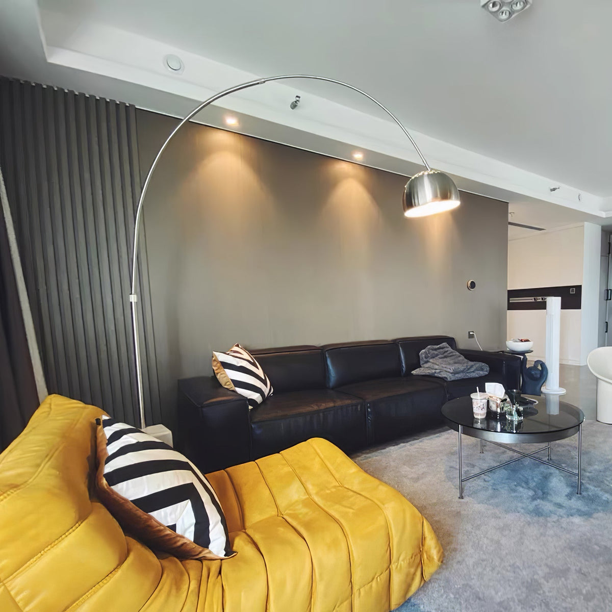 In a minimalist white room, the Clérachic arc lamp's marble base stands beside a black leather three-seat sofa on stone flooring.