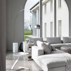 Through a white archway, the Clérachic arc lamp's marble base stands beside a pale grey sectional sofa with a garden view.