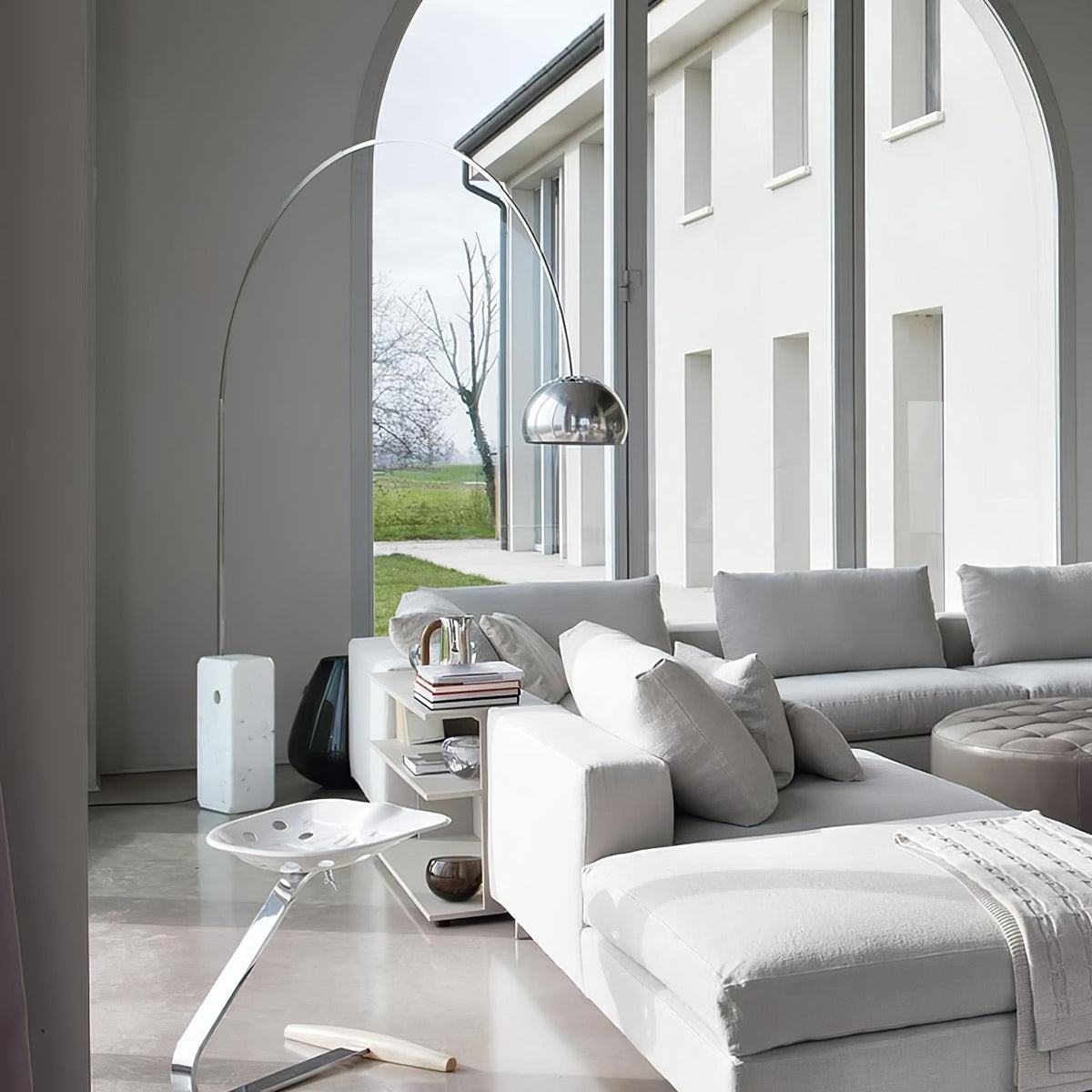 Through a white archway, the Clérachic arc lamp's marble base stands beside a pale grey sectional sofa with a garden view.