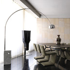 The Clérachic arc lamp's chrome dome hangs above a dark wood dining table with taupe chairs behind vertical blinds.