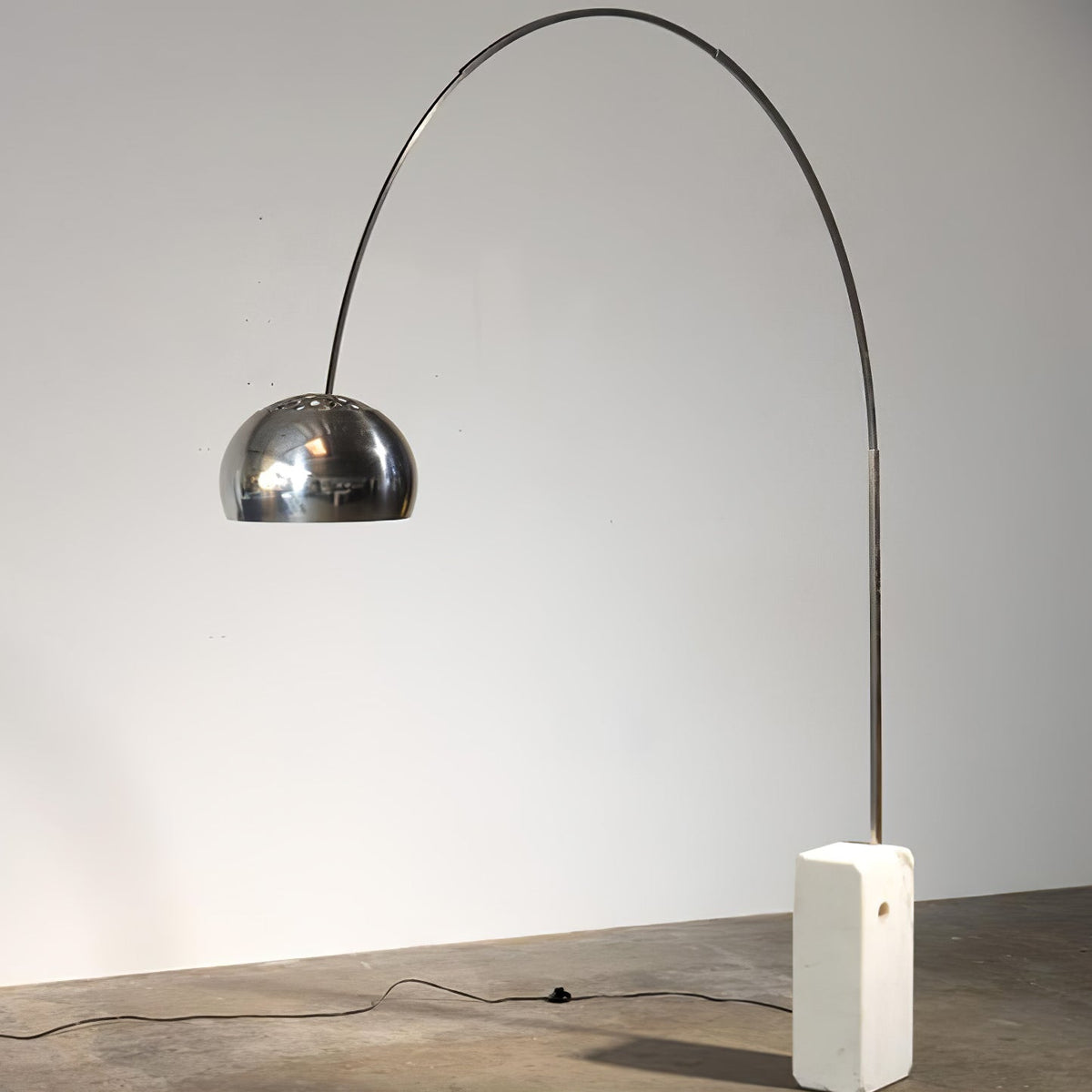 A Clérachic arc floor lamp stands on a plain grey backdrop, its power cord visible trailing across the studio floor.