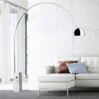 The Clérachic arc lamp's chrome dome curves above a white leather sectional sofa with rust and blue striped cushions.