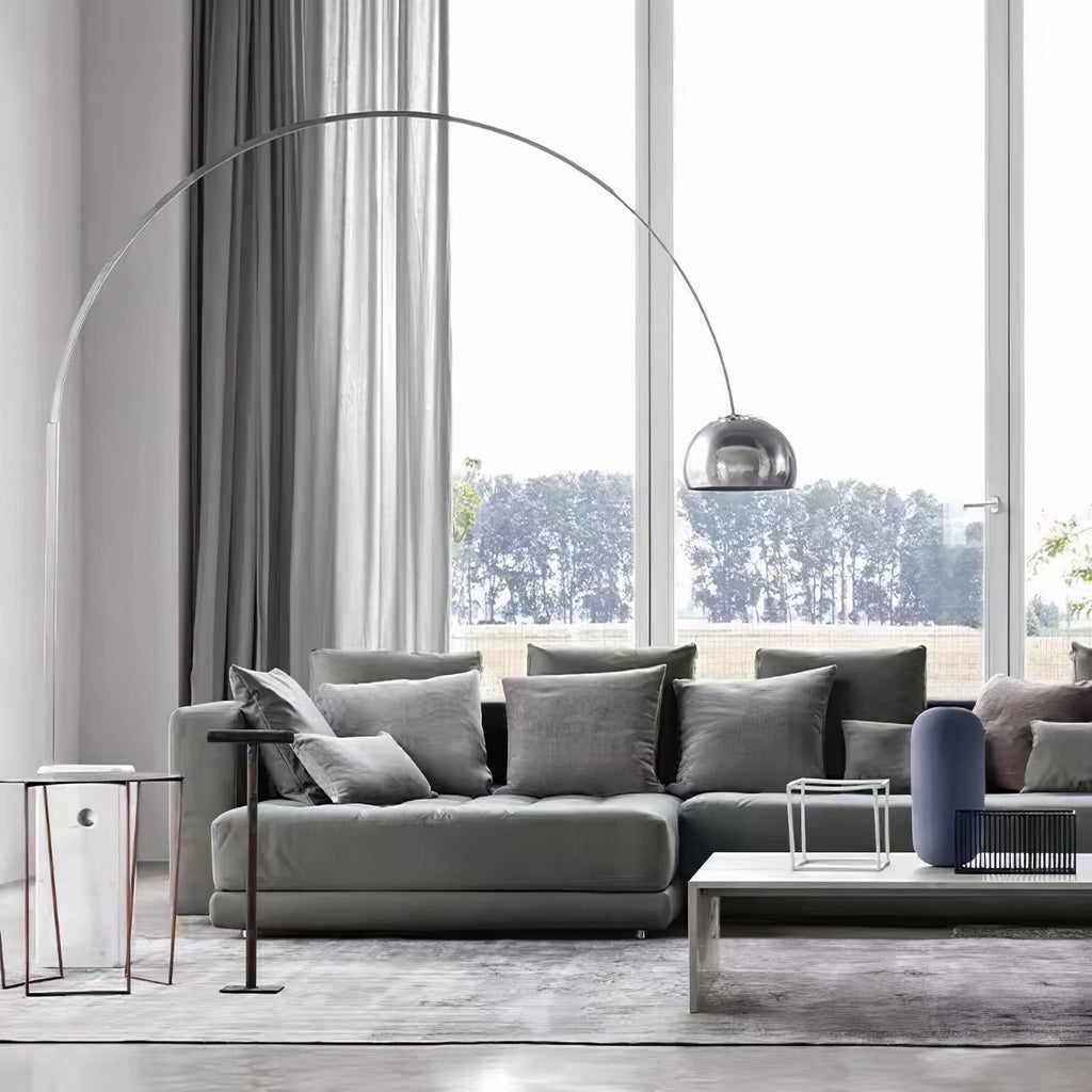 The Clérachic arc lamp's chrome dome curves above a grey sectional sofa facing tall windows that look onto a green lawn.
