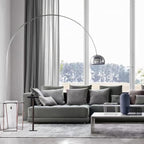 The Clérachic arc lamp's chrome dome curves above a grey sectional sofa facing tall windows that look onto a green lawn.