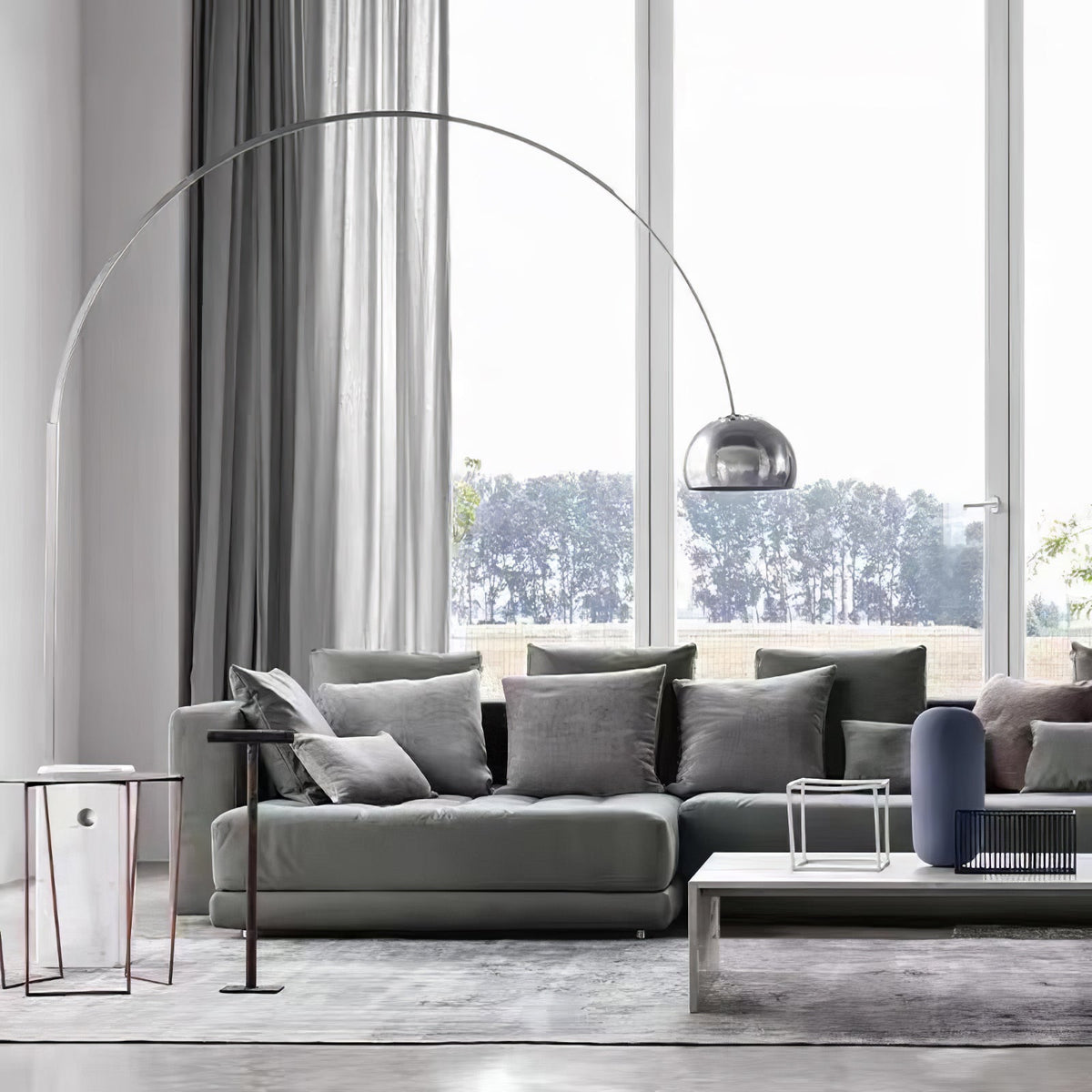 The Clérachic arc lamp's chrome dome curves above a grey sectional sofa facing tall windows that look onto a green lawn.