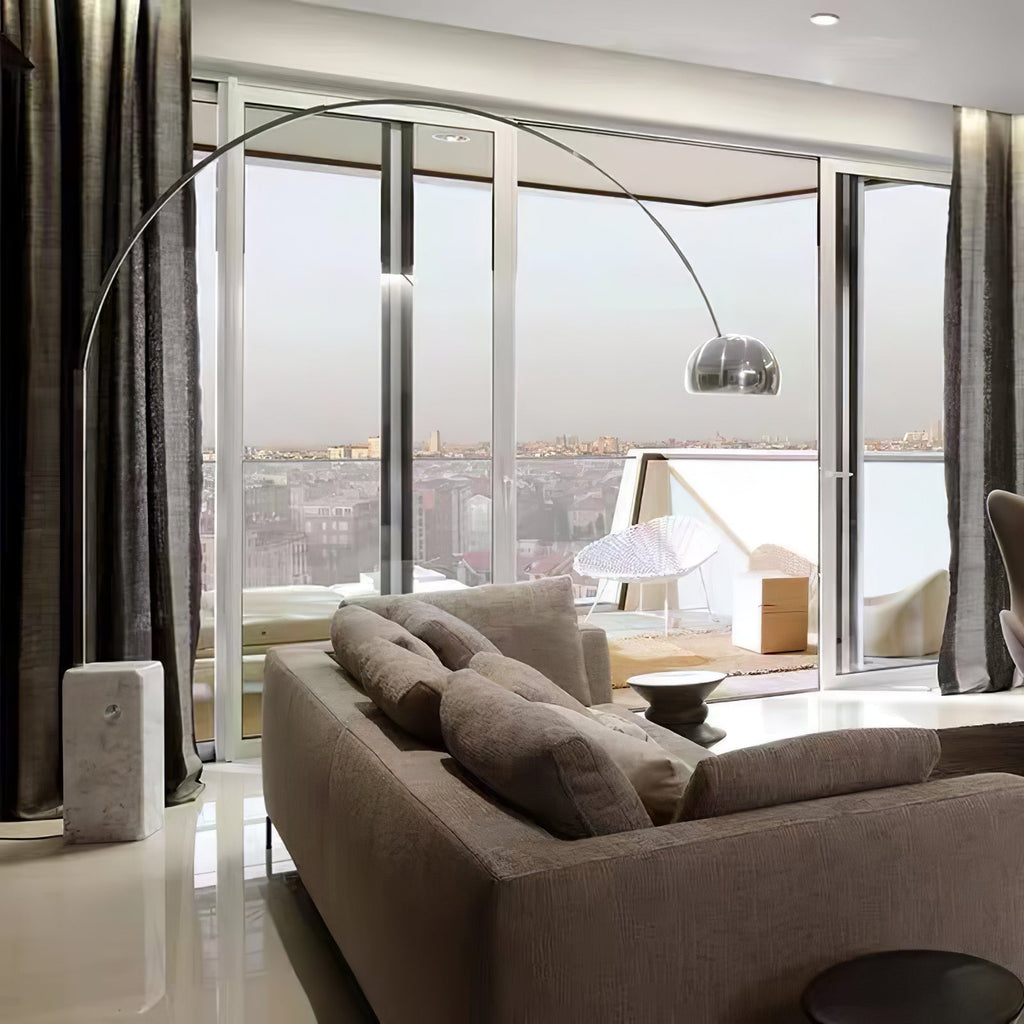 A Clérachic arc lamp's marble base stands beside a dark sectional sofa, chrome shade catching a floor-to-ceiling city skyline view.