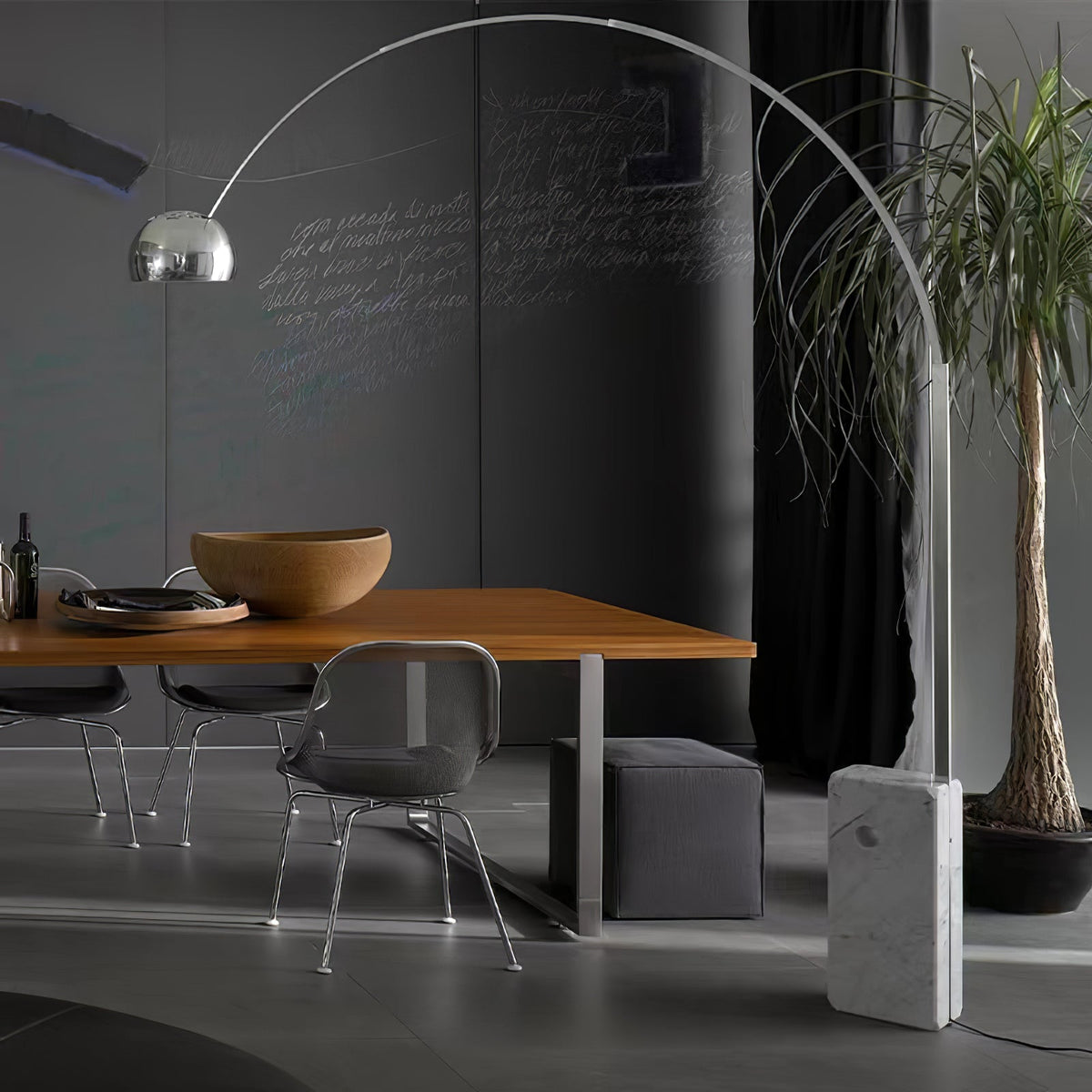 In a dark chalkboard-walled dining room, the Clérachic arc lamp's marble base sits beside a wooden table and potted palm.