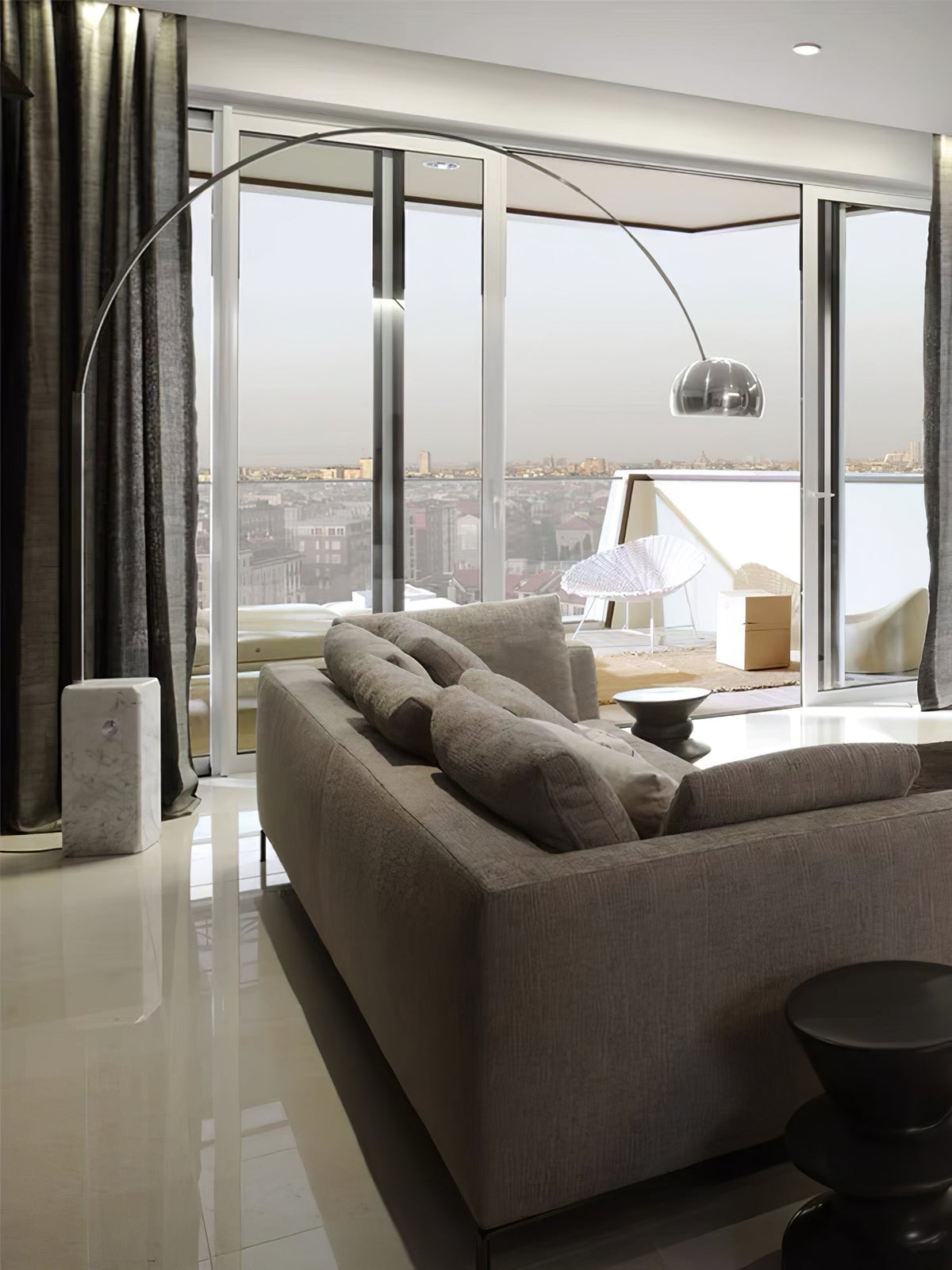From a high-rise lounge, the Clérachic arc lamp's chrome dome frames a rooftop terrace and city skyline beyond the sofa.