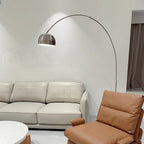 The Clérachic arc lamp's lit chrome dome glows above a cream leather sofa and tan leather armchair by a terrazzo table.