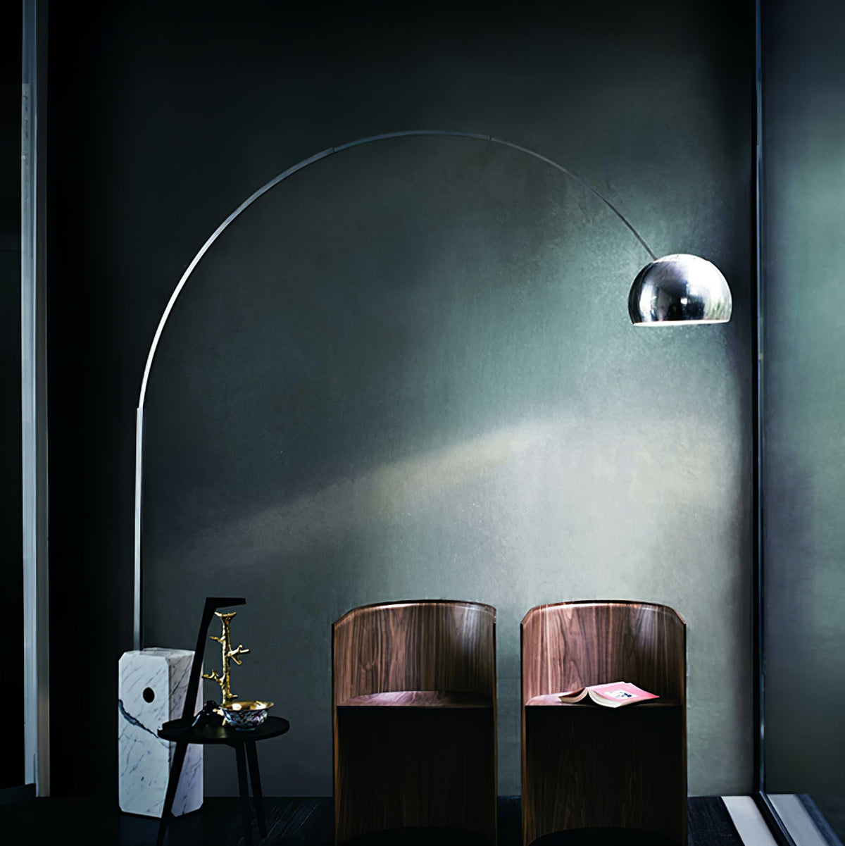 In a moody dark room, the Clérachic arc lamp's lit dome illuminates two curved walnut chairs and a small brass-topped table.