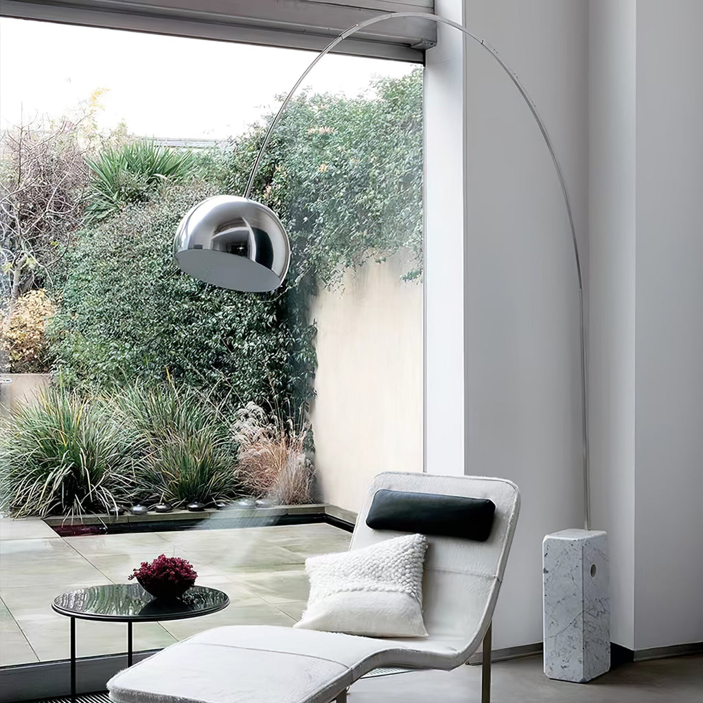 The Clérachic arc lamp's marble base sits beside a white bouclé chaise, its chrome dome framed by a dense garden view.