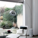 The Clérachic arc lamp's marble base sits beside a white bouclé chaise, its chrome dome framed by a dense garden view.