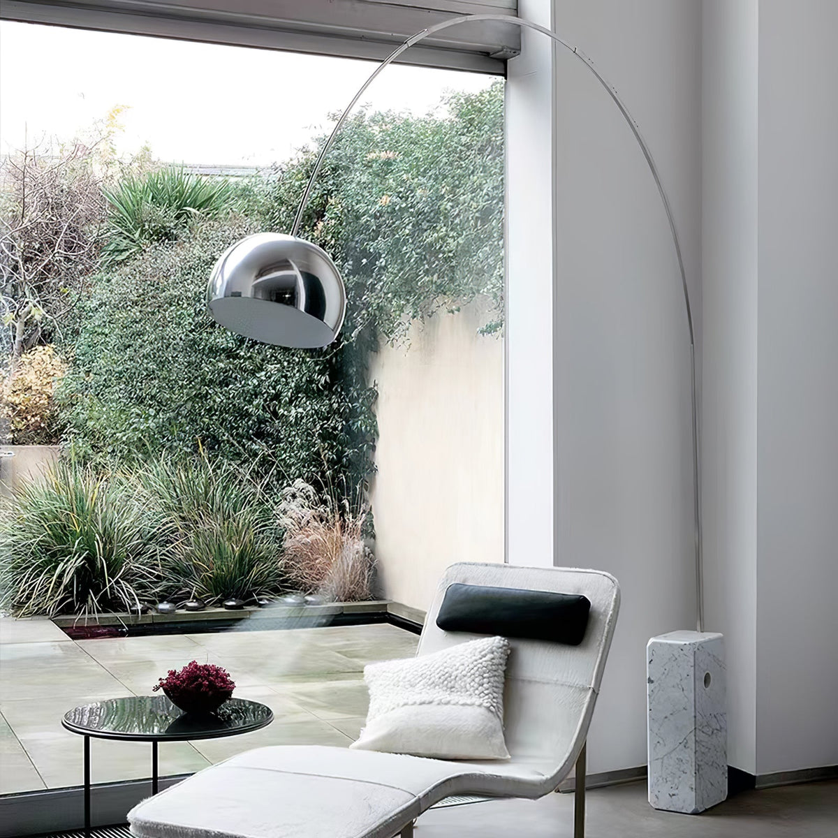 The Clérachic arc lamp's marble base sits beside a white bouclé chaise, its chrome dome framed by a dense garden view.