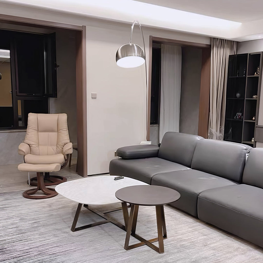 In an open-plan flat, the Clérachic arc lamp curves above a grey sectional sofa, a cream recliner chair visible across the room.
