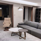 In an open-plan flat, the Clérachic arc lamp curves above a grey sectional sofa, a cream recliner chair visible across the room.