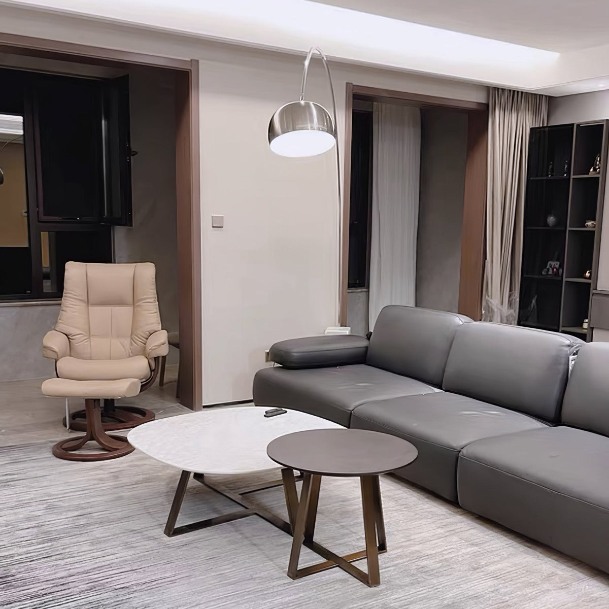 In an open-plan flat, the Clérachic arc lamp curves above a grey sectional sofa, a cream recliner chair visible across the room.