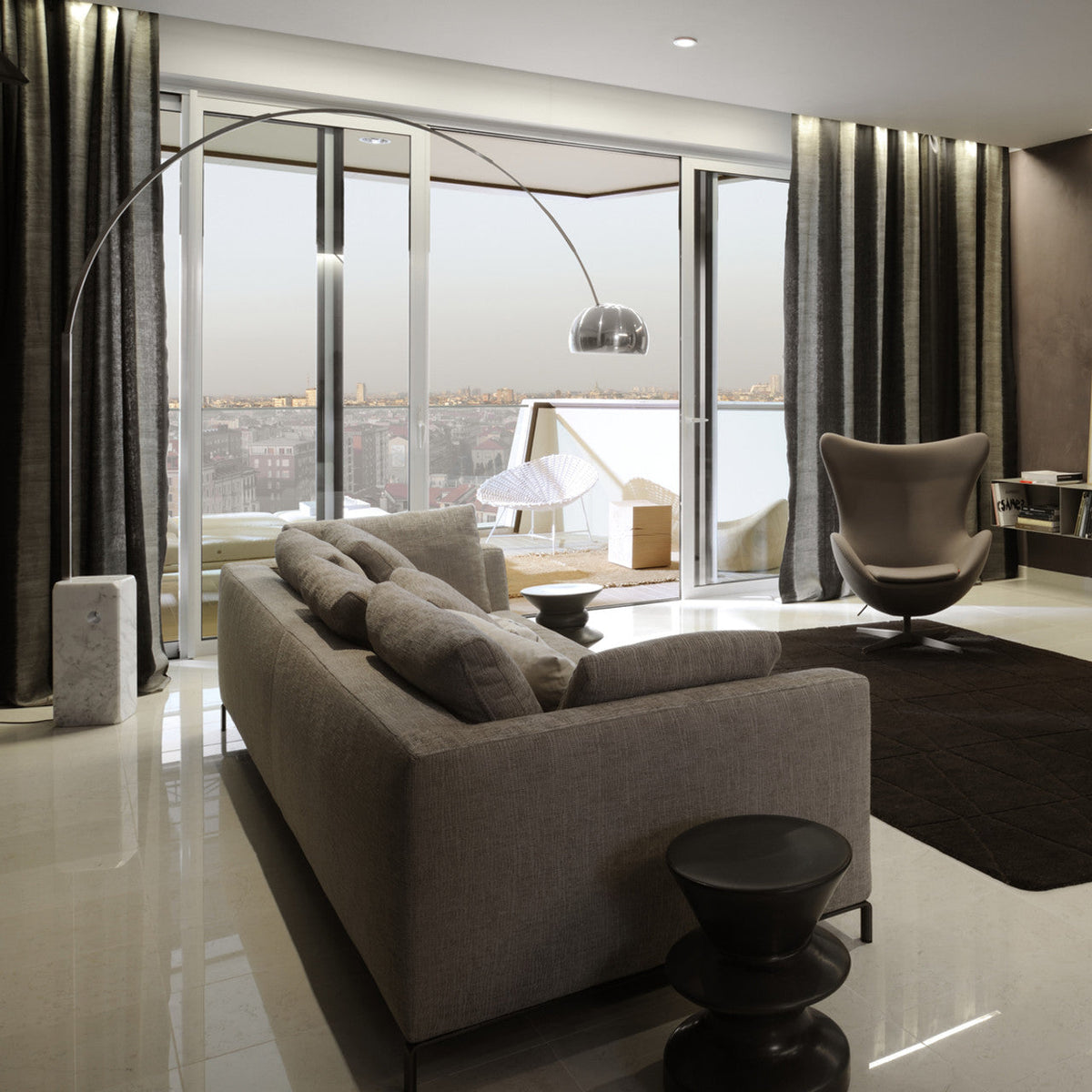 At dusk, the Clérachic arc lamp's chrome dome glows softly over a grey sofa in a high-rise flat with a city skyline behind it.
