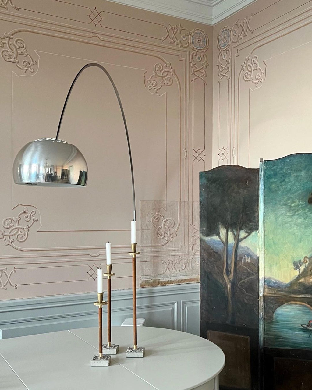 In a rose-pink panelled room, the Clérachic lamp's slim steel arch curves above brass candlesticks and a painted folding screen.