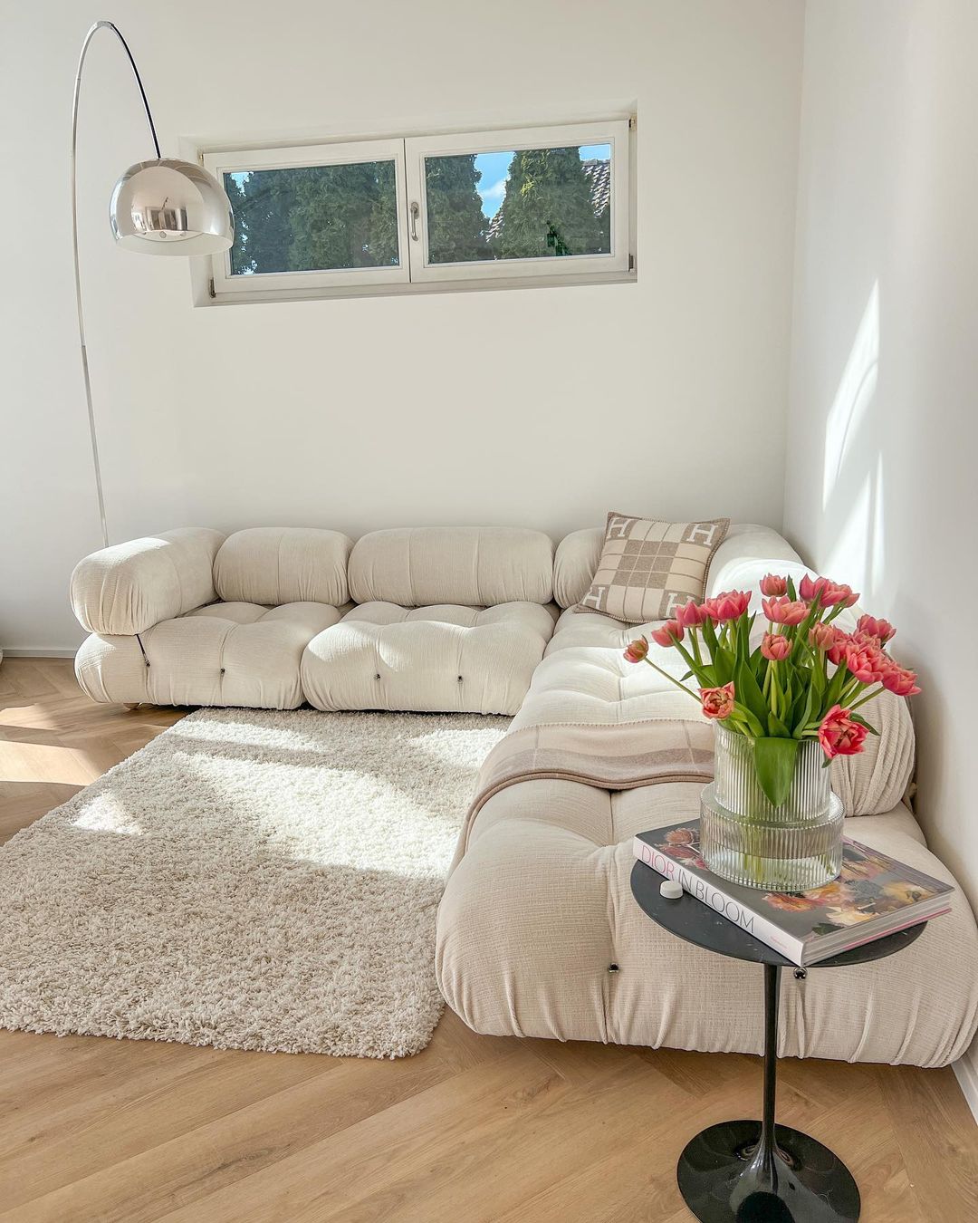 The Clérachic arc lamp's marble base anchors a bright corner beside a cream tufted sofa and a glass vase of pink tulips.
