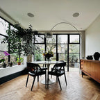 The Clérachic arc lamp curves above a marble dining table and black leather chairs, framed by tall steel-mullioned windows.