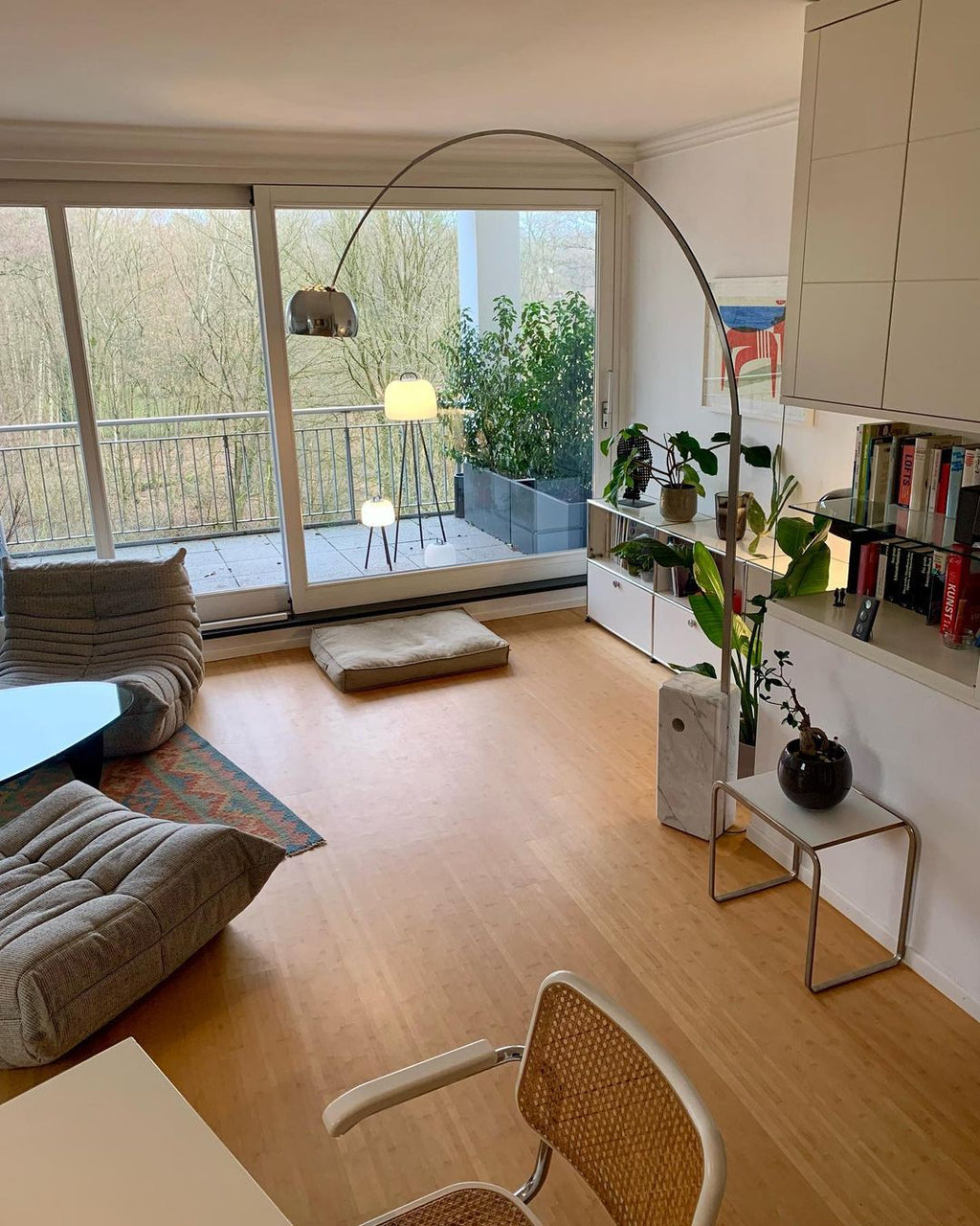 The Clérachic arc lamp's chrome dome hangs above a floor cushion and low sofa, with a balcony and bare trees visible beyond.