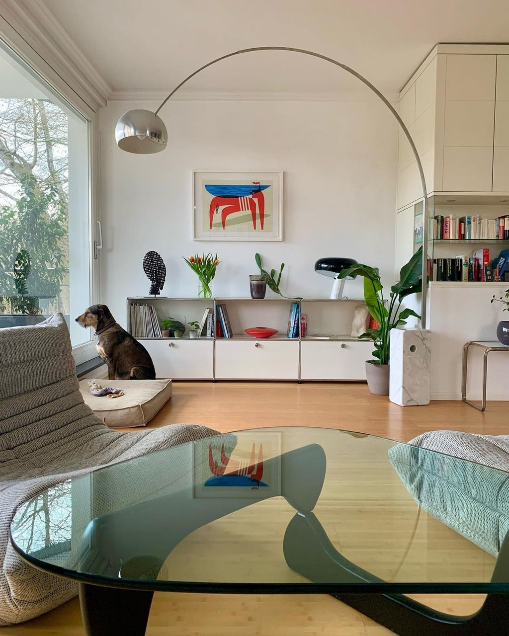 A dog rests on its bed beneath the Clérachic arc lamp, its chrome dome reflected in a glass coffee table across the room.