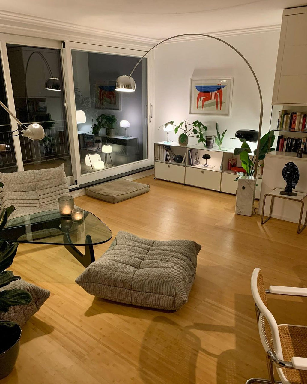 At night, the Clérachic arc lamp's chrome dome glows among reflected lamps in a glass door, lighting a cosy seating area.