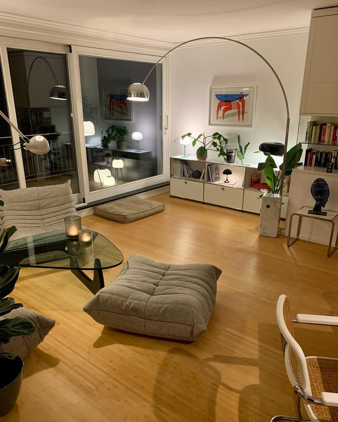 At night, the Clérachic arc lamp's chrome dome glows among reflected lamps in a glass door, lighting a cosy seating area.