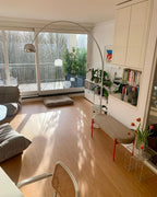 Sunlight streaks across the floor as the Clérachic arc lamp curves above a low bench with tulips near a balcony doorway.