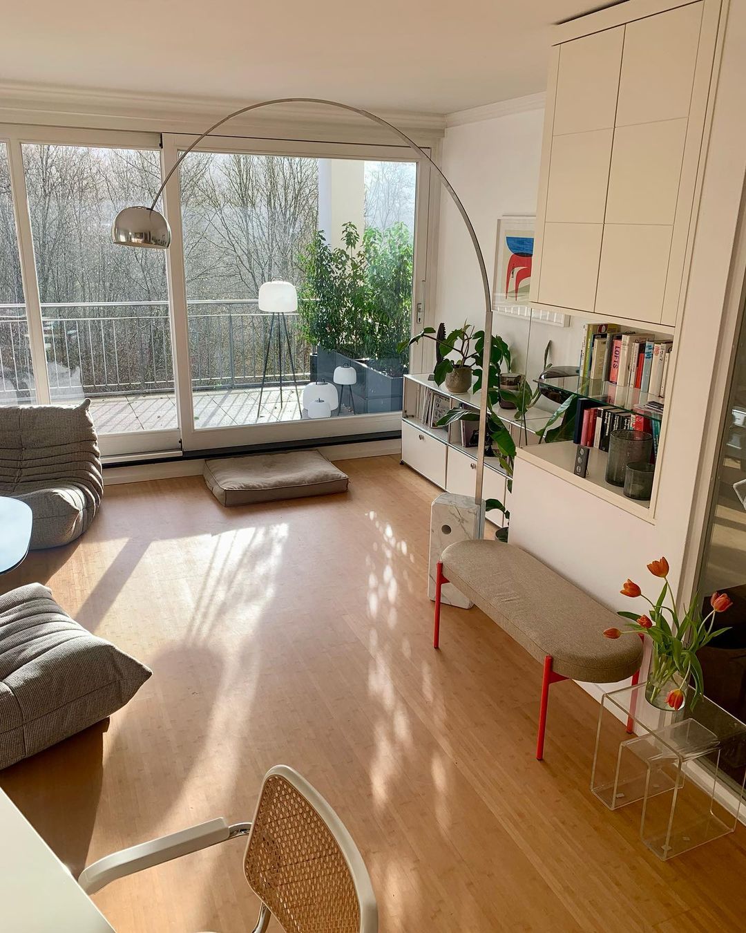 Sunlight streaks across the floor as the Clérachic arc lamp curves above a low bench with tulips near a balcony doorway.