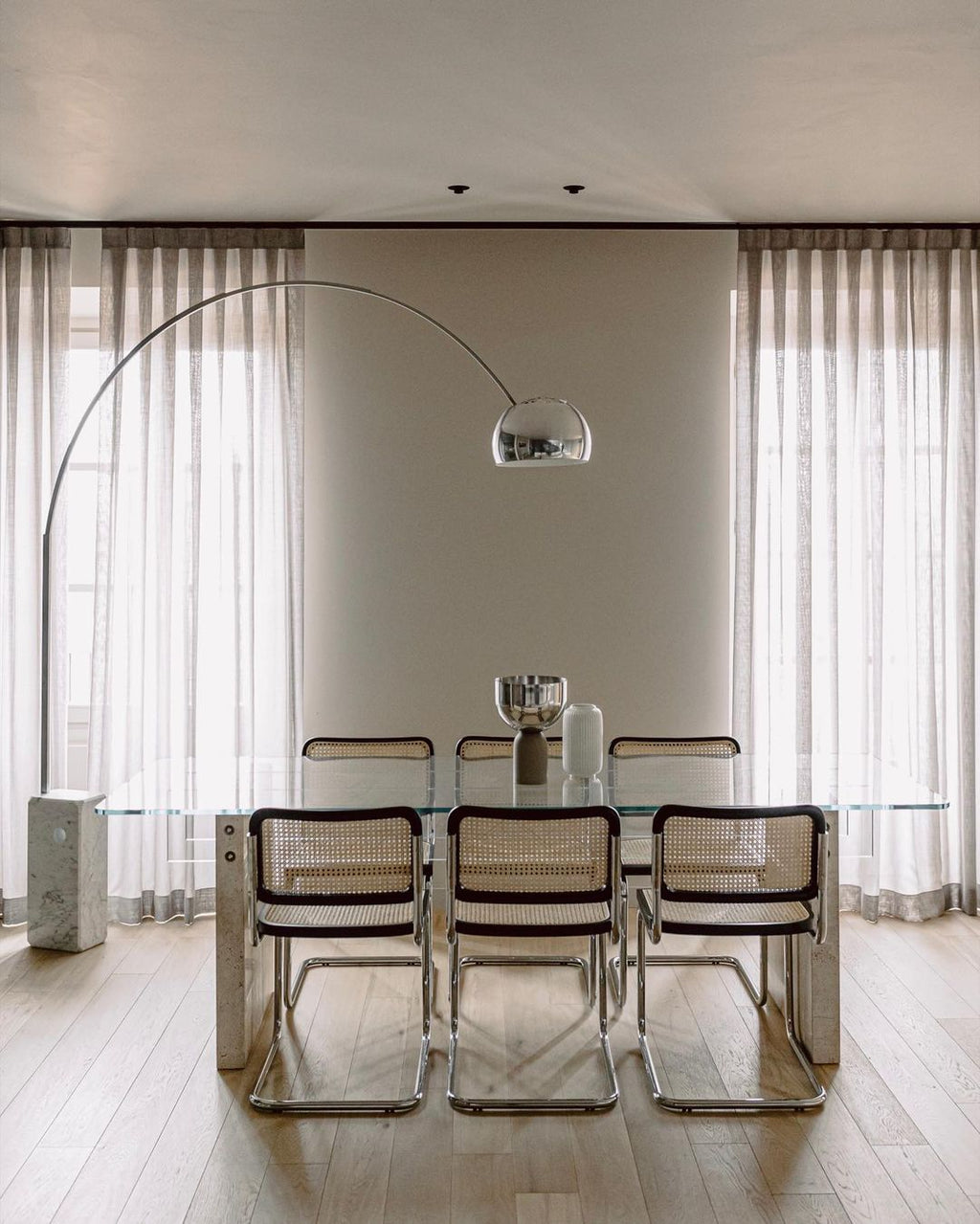 Clérachic's arc lamp curves above a glass-topped dining table with cane-backed chrome chairs and sheer floor-length curtains.