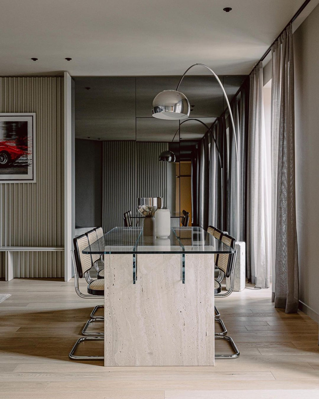 Reflected in a mirrored wall, the Clérachic arc lamp's dome hangs above a travertine-based dining table with a glass top.