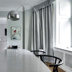 The Clérachic arc lamp's chrome dome hangs low over a pale dining table, its marble base framed by soft grey curtains.